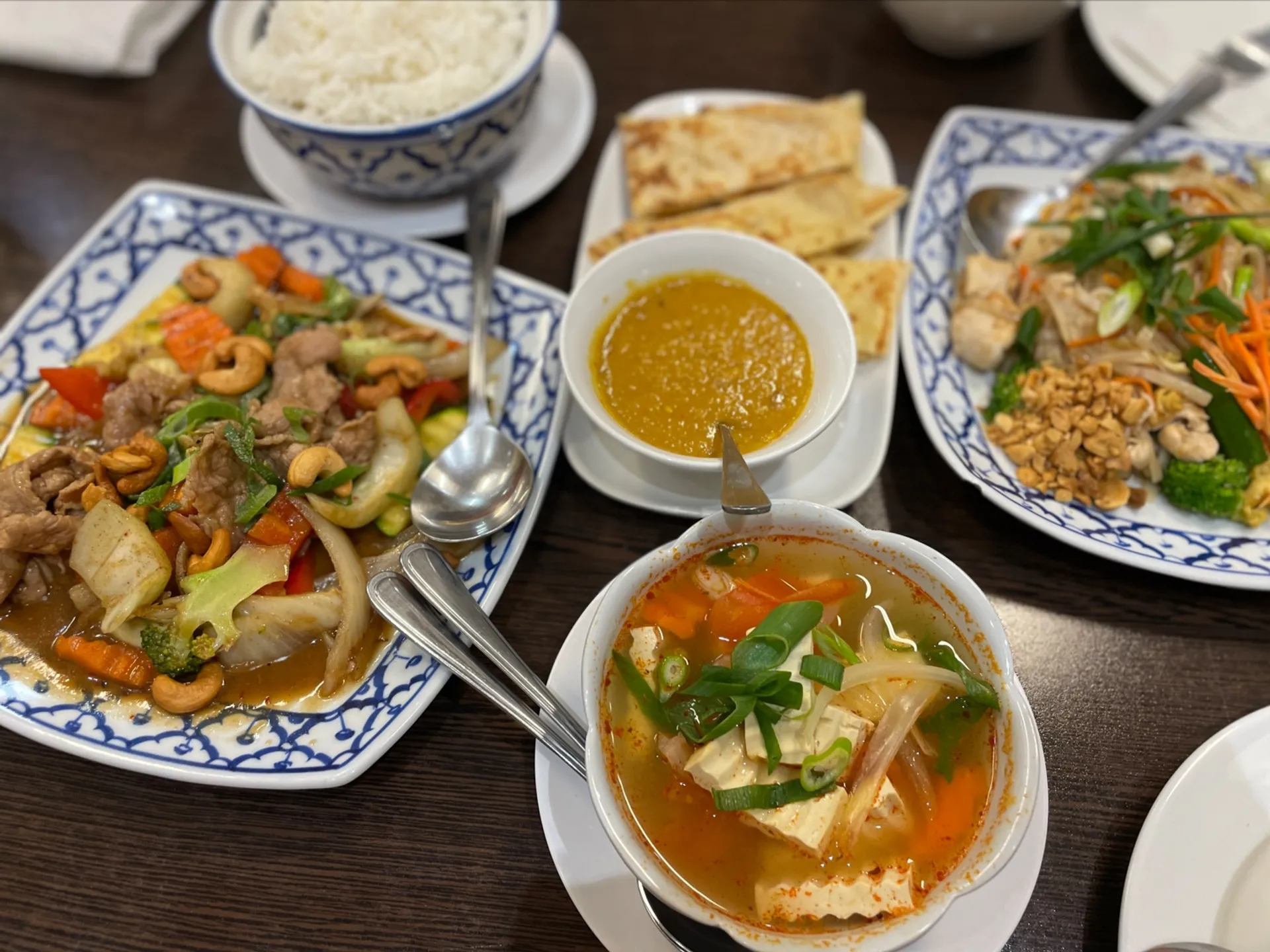 Table spread of four Thai dishes including stir-fry, soup, roti and noodles on blue-white ceramic plates
