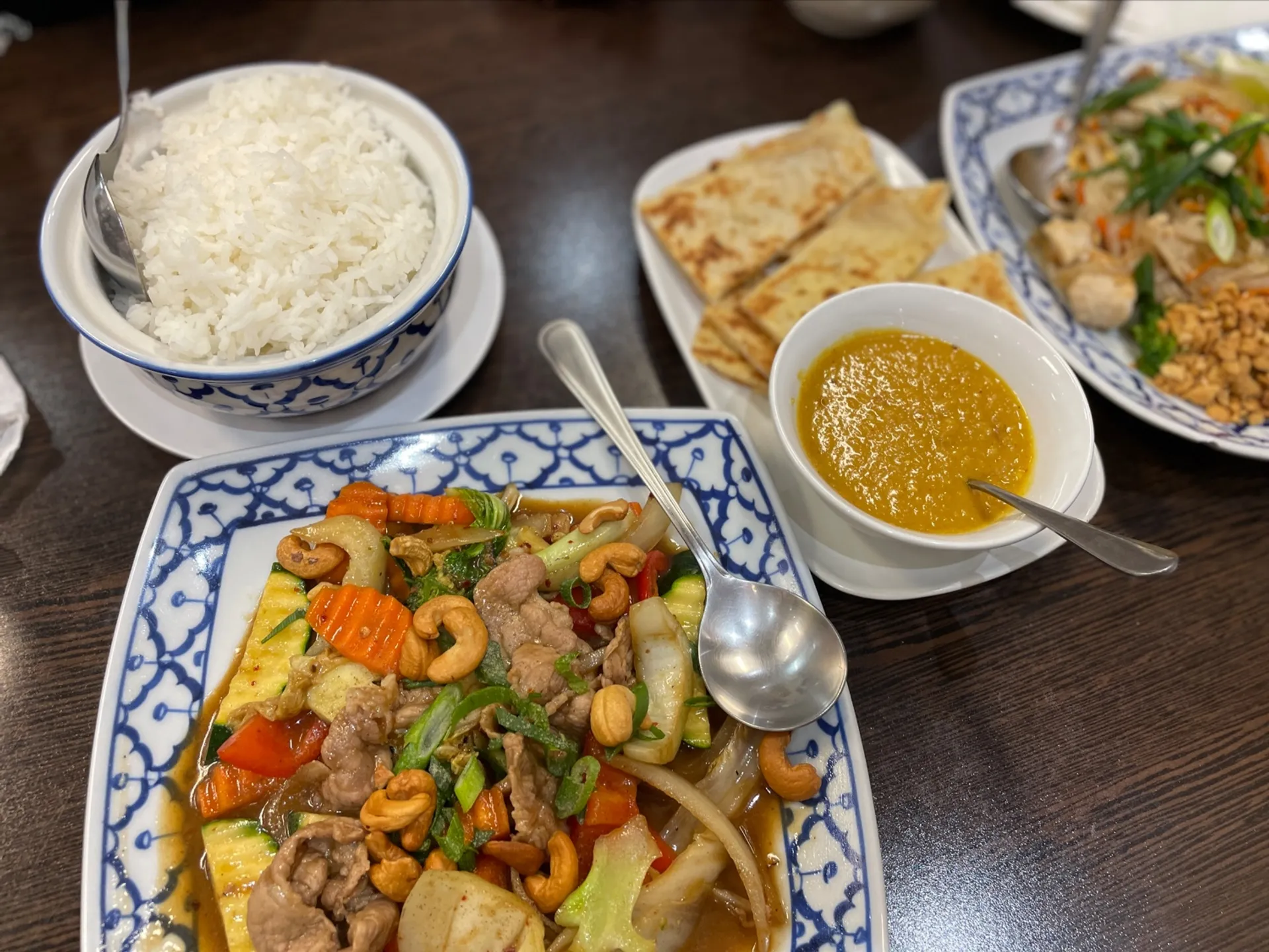 Table spread of Thai dishes including stir-fry, rice, roti and satay sauce on blue-white ceramic plates